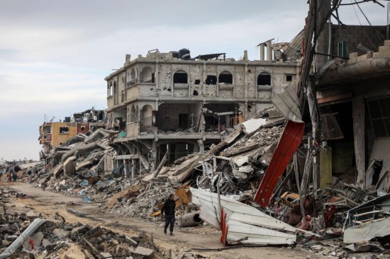 2月9日，在加沙地帶內(nèi)察里姆走廊附近，巴勒斯坦人經(jīng)過(guò)建筑廢墟。新華社發(fā)（里澤克·阿卜杜勒賈瓦德攝）